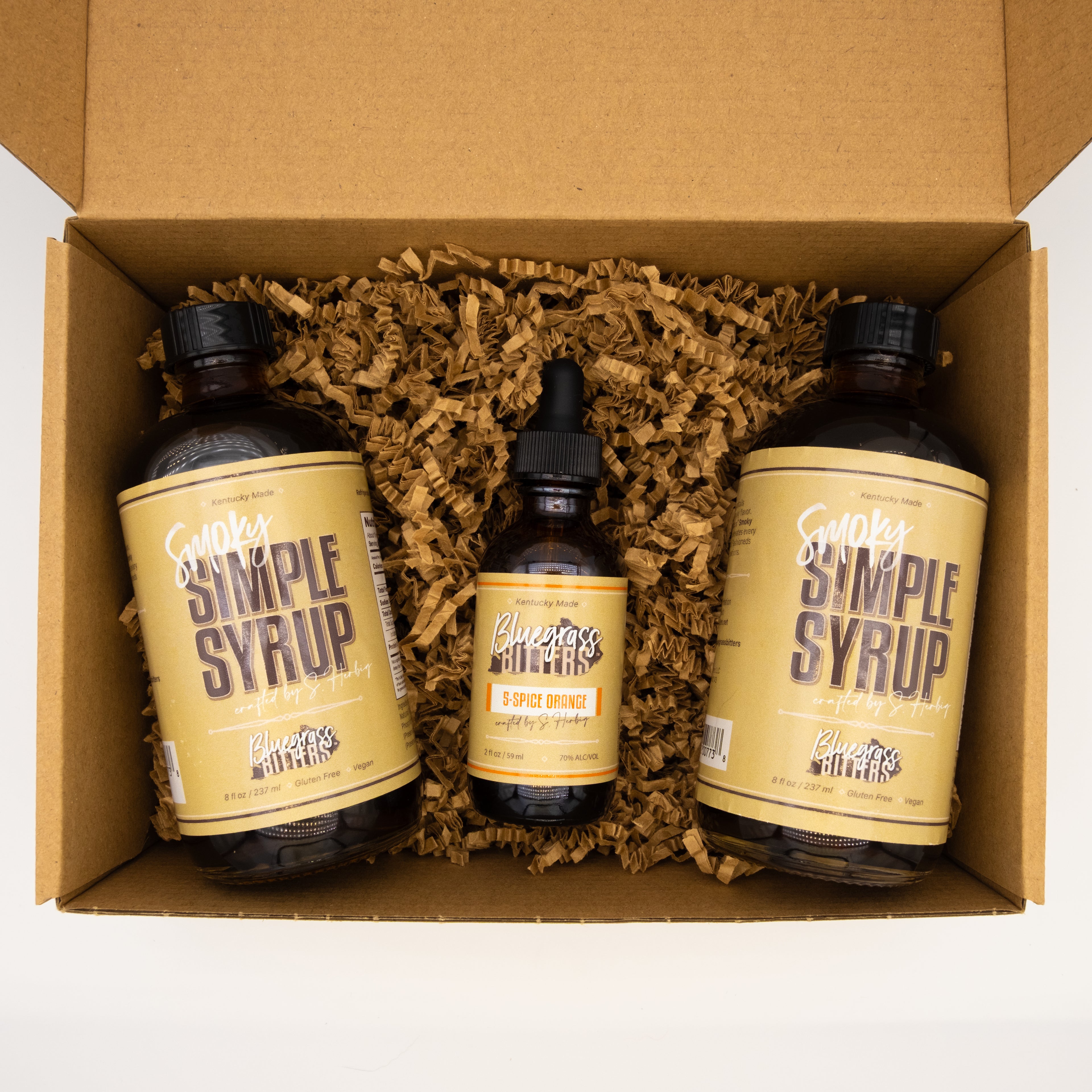 Two bottles of Smoky Simple Syrup and one bottle of 5-Spice Orange Bitters in a cardboard box with brown filler