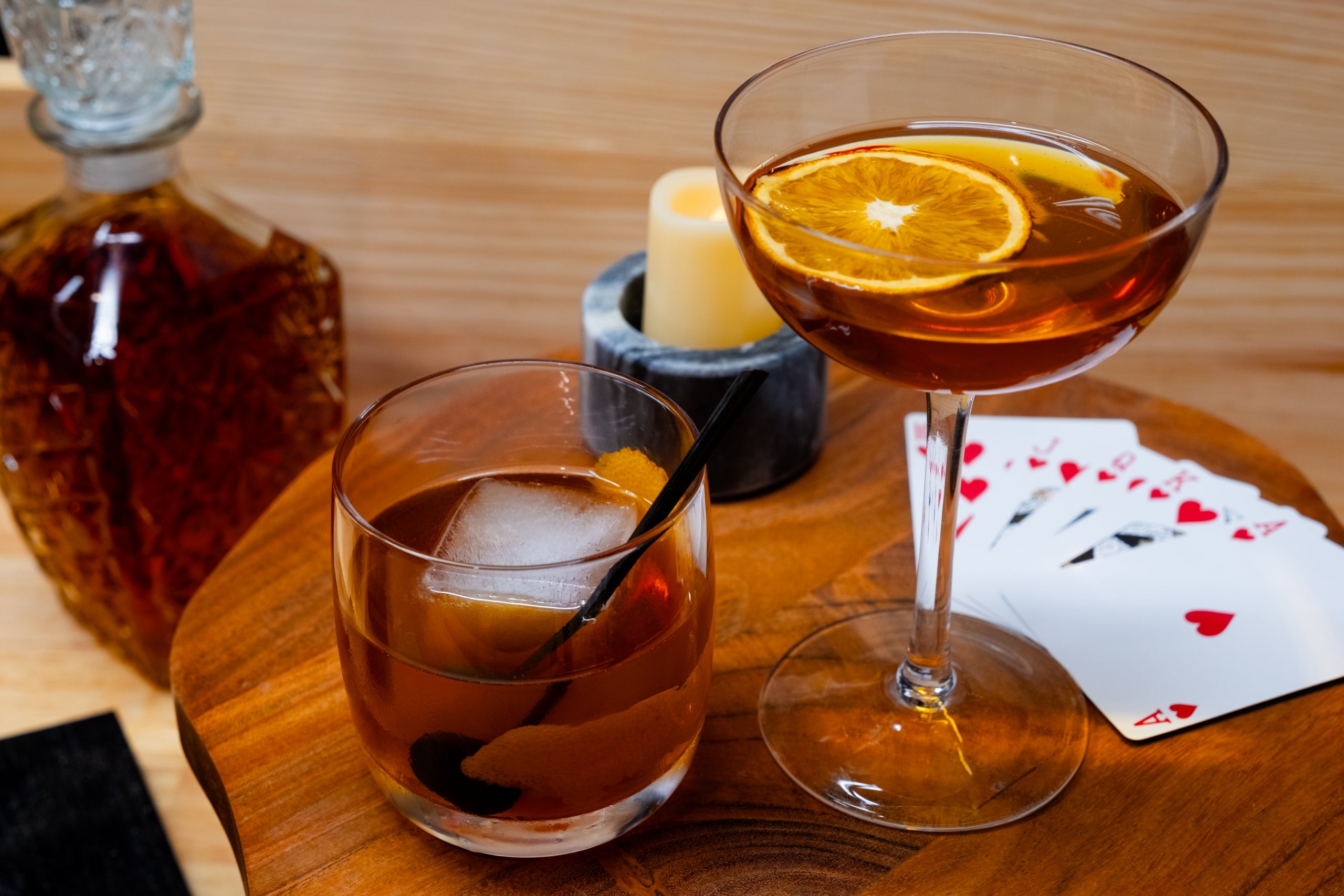 Two cocktails with ice and orange slices on a wooden tray with playing cards and a bottle of whiskey.
