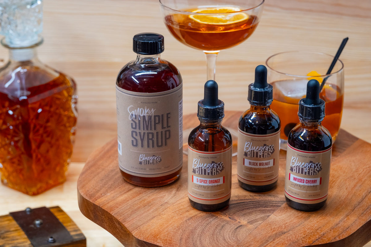 Bluegrass Bitters Smoky Simple Syrup and bitters bottles on a wooden tray with cocktails.