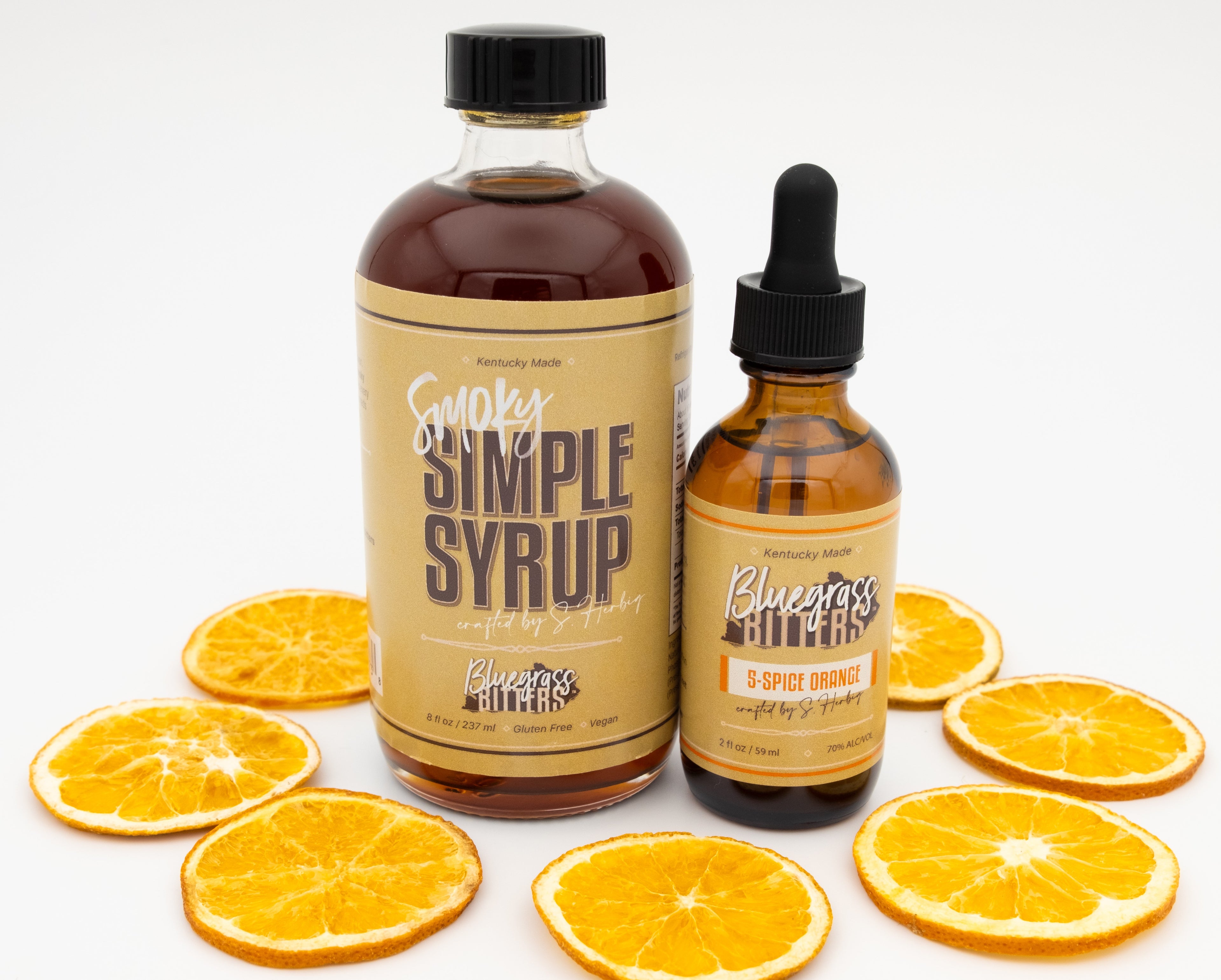 One bottle of Smoky Simple Syrup and one bottle of 5-Spice Orange bitters with orange slices on a white background