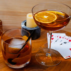 Two cocktails with orange slices and ice on a wooden table with playing cards.