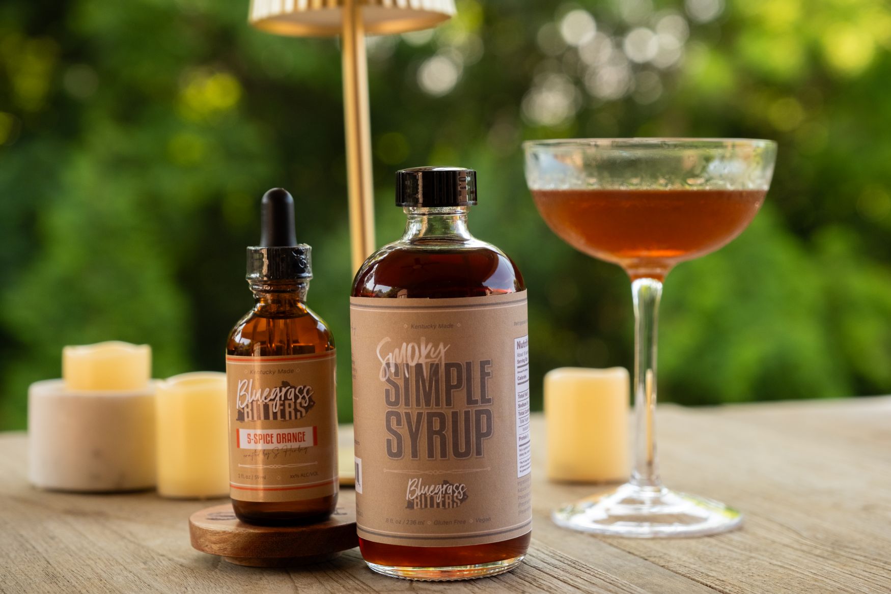 A bottle of Bluegrass Bitters 5-Spice Orange Bitters and Smoky Simple Syrup sit on an outdoor table. A Burnt Orange cocktail and decorative props are in the background.